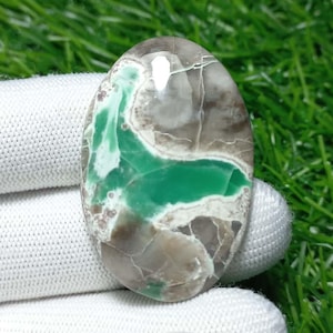 Puede incluir: Una gema ovalada con una superficie pulida. La piedra presenta un centro verde vibrante, rodeado de áreas grises, marrones y blancas. La gema se sostiene sobre un fondo de tela blanca y hierba verde.