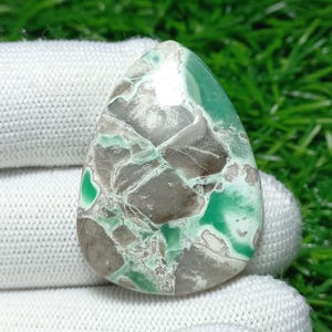 Puede incluir: Una gema en forma de lágrima con un patrón único de tonos grises, marrones y verdes. La superficie pulida revela un diseño en forma de mosaico, con áreas de verde vibrante y una mezcla de tonos terrosos. La piedra se sostiene contra una tela blanca.