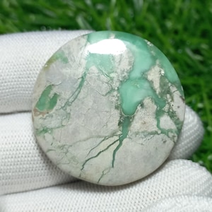 Puede incluir: Una gema redonda y pulida, con una mezcla de colores verde claro y blanco. La piedra tiene un aspecto veteado con vetas de verde más oscuro. La superficie es lisa y reflectante, con un ligero brillo.