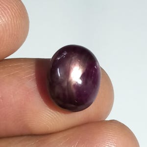 May include: A deep purple, oval gemstone with a star effect. The polished stone is held between fingers, showing its shape and colour. The gem has a bright, reflective centre against a white background. The stone's shape is oval.