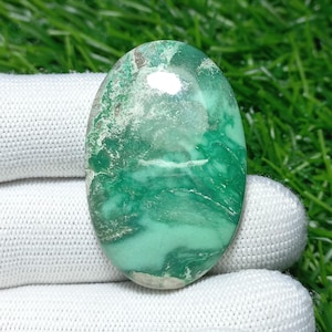 Puede incluir: Gema ovalada con superficie pulida. La piedra presenta una mezcla de tonos verdes, con tonos más claros arremolinándose a través de una base más oscura. La gema se encuentra sobre un tejido blanco y un fondo de hierba verde.