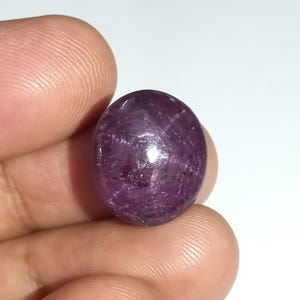 May include: A polished, oval-shaped, purplish-red gemstone. The gem has a smooth, reflective surface with subtle internal lines. The stone is held between fingers against a white background, highlighting its colour and shape. This gemstone is likely a ruby.