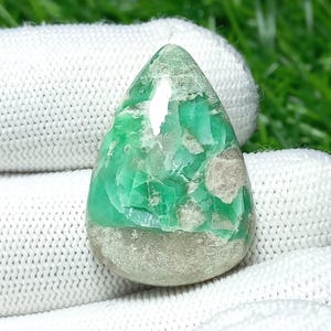 Puede incluir: Una gema en forma de lágrima, de color verde vibrante, con áreas grises claras y una base texturizada gris claro. La superficie pulida refleja la luz, resaltando los patrones naturales de la piedra. La gema se encuentra sobre una tela blanca.