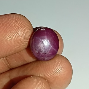 Könnte beinhalten: Ein polierter, ovalförmiger Rubin-Edelstein. Der Edelstein hat eine tiefe, satte rote Farbe mit subtilen inneren Mustern. Der Stein wird zwischen Fingern gehalten, was seine glatte, reflektierende Oberfläche zeigt. Geeignet für die Schmuckherstellung.