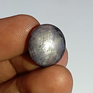 Puede incluir: Una piedra preciosa de zafiro estrella de forma redonda y corte cabujón, con un tono grisáceo-púrpura translúcido. La superficie muestra un efecto de asterismo de seis rayos, formado por líneas de luz que crean un patrón de estrella.