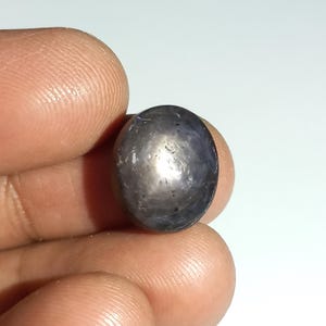 Può includere: Una gemma cabochon di forma ovale color blu scuro caratterizzata da un distinto effetto a stella a sei raggi noto come asterismo. La superficie della pietra è liscia e lucida, mostrando una tonalità blu profonda e satura con un riflesso di luce centrato.
