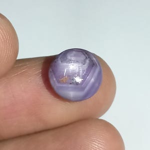 May include: A faceted, lavender-colored gemstone with a rounded shape. The gem has a complex, geometric pattern of facets and appears to be translucent. The stone is held between fingers, showcasing its size and color.
