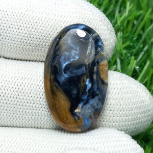 May include: An oval-shaped Pietersite gemstone with a polished surface. The stone features swirling patterns of deep blue, gold, and gray hues. The gemstone is held against a white fabric background and green grass.