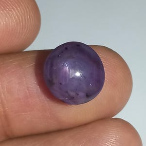 May include: A round, polished gemstone with a deep purple hue. The stone has a smooth surface with subtle variations in color and small dark inclusions. The gem is held between two fingers against a plain white background.