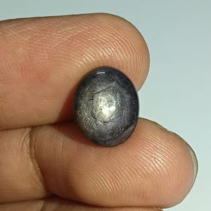 Könnte beinhalten: Ein dunkler, ovaler Edelstein mit sternförmigem Muster. Die polierte Oberfläche des Steins reflektiert das Licht. Der Edelstein wird zwischen Fingern gehalten.