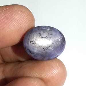 Puede incluir: Una piedra preciosa en forma de cabujón ovalado con un tono púrpura claro translúcido. La piedra presenta un efecto de estrella de seis puntas en su superficie pulida, junto con inclusiones oscuras e irregulares dispersas en su interior.
