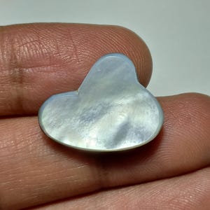 May include: A pearlescent cloud-shaped gemstone with a light blue and white iridescent surface. The gemstone is held in a person's hand, showcasing its smooth, polished finish. The cloud shape is rounded and symmetrical, with a subtle sheen.