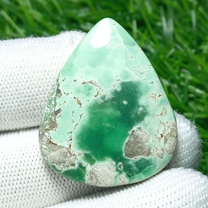 Puede incluir: Una gema en forma de lágrima con una superficie pulida. La piedra presenta una mezcla de tonos verdes claros y oscuros, con áreas de color beige y gris. La gema se sostiene sobre un fondo de tela blanca y hierba verde.