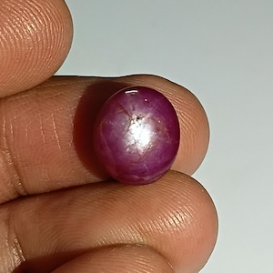 Könnte beinhalten: Ein polierter, ovaler Rubin-Edelstein. Der Edelstein ist ein tiefes, sattes Rot mit einem sternartigen Effekt auf der Oberfläche. Der Stein wird in einer Hand gehalten, um seine Größe und Farbe zu zeigen. Dieser Edelstein eignet sich für die Schmuckherstellung.