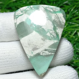 Puede incluir: Una gema triangular con una mezcla de colores, incluyendo blanco, gris y tonos verdes. La piedra tiene una superficie pulida con un patrón único de vetas y remolinos. La gema se sostiene en un guante blanco.