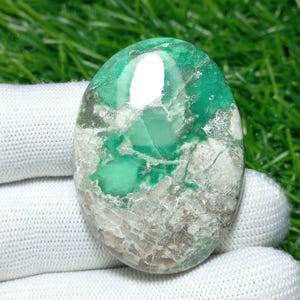 Puede incluir: Gema ovalada con una mezcla de tonos verdes, blancos y grises. La superficie pulida refleja la luz, resaltando los patrones y texturas naturales. El color de la piedra varía de verde claro a oscuro, con áreas blancas y grises.