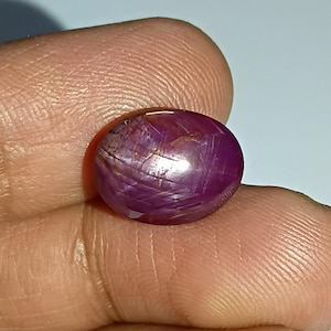 Könnte beinhalten: Ein polierter, ovalförmiger Rubin-Edelstein. Der Edelstein hat eine tiefe, satte rot-violette Farbe mit sichtbaren inneren Streifen. Der Stein wird zwischen den Fingern gehalten und zeigt seine glatte, reflektierende Oberfläche. Dieser Edelstein eignet sich für die Schmuckherstellung.