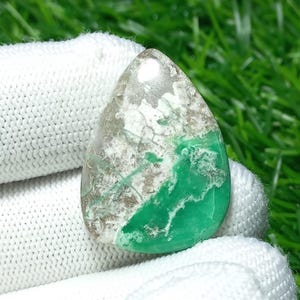 Puede incluir: Una piedra preciosa en forma de lágrima con una mezcla de colores. La mitad superior es un gris claro translúcido con patrones blancos y marrones. La mitad inferior es un verde vibrante con vetas blancas. La piedra se sostiene en un guante blanco.