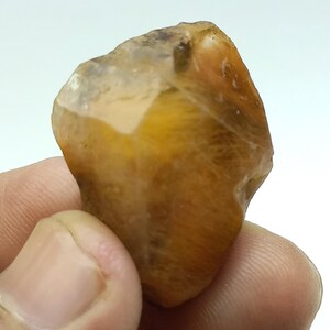 May include: A natural gemstone in shades of golden yellow and translucent white. The stone has a textured surface and an irregular shape, highlighting its raw, unpolished appearance. The gemstone is held in a hand.
