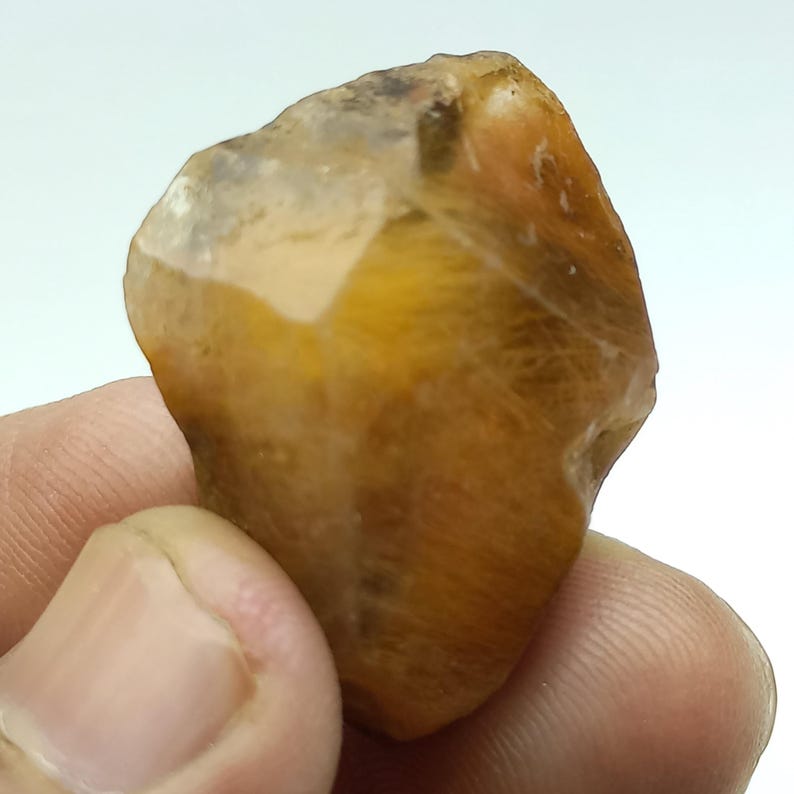 May include: A natural gemstone in shades of golden yellow and translucent white. The stone has a textured surface and an irregular shape, highlighting its raw, unpolished appearance. The gemstone is held in a hand.