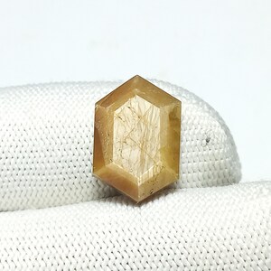 May include: A golden-brown, hexagonal gemstone with multiple facets. The gem has internal inclusions and a polished surface. The shape is a modified hexagon, with multiple facets creating a sparkling effect. The gemstone is set against a white background.