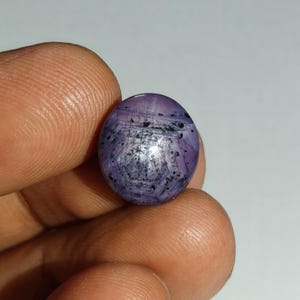 Può includere: Una gemma viola di forma rotonda e taglio cabochon che presenta un effetto a stella a sei punte noto come asterismo. La pietra mostra una tonalità viola traslucida con inclusioni minerali scure sparse e un distinto motivo a griglia geometrica.