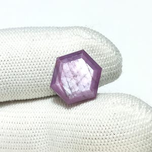 May include: A hexagonal cut pink sapphire gemstone with a faceted design. The gemstone is set against a white fabric background.