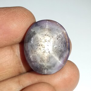 Può includere: Una gemma a cabochon di forma ovale caratterizzata da un evidente effetto a stella a sei punte, noto come asterismo. La pietra presenta una tonalità traslucida viola chiaro e bianca con inclusioni interne visibili e una superficie liscia e lucida.