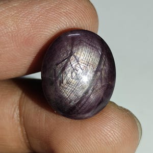 May include: A polished, oval-shaped gemstone with a deep purple hue. The stone features a shimmering, star-like effect with silver and gold-colored inclusions. The gemstone is held between two fingers, showcasing its size and color.
