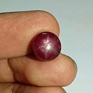 Könnte beinhalten: Ein runder, polierter Rubin-Edelstein mit Sterneneffekt. Der Edelstein hat eine tiefrote Farbe mit einem sechsstrahligen Sternmuster auf der Oberfläche. Der Rubin wird zwischen Fingern gehalten, um seine Größe und Farbe zu zeigen.