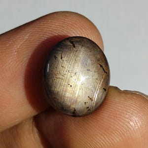 Puede incluir: Una piedra preciosa cabujón pulida de forma ovalada con una superficie de color bronce metálico y marrón oscuro, con inclusiones lineales visibles y un reflejo de luz chatoyante distintivo que recorre verticalmente el centro de la cúpula lisa.