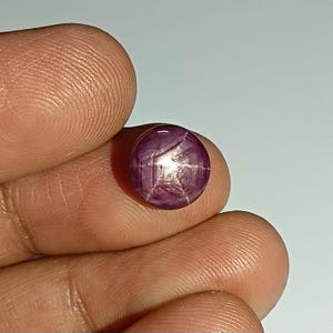 Könnte beinhalten: Ein runder, polierter Sternrubin-Edelstein. Der Edelstein hat eine tiefrote bis violette Farbe und zeigt ein sechsstrahliges Sternmuster auf seiner Oberfläche. Der Stein wird zwischen Fingern vor hellem Hintergrund gehalten.
