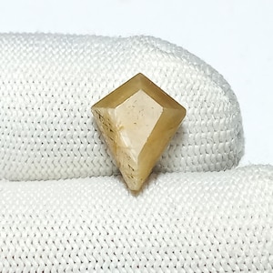 May include: A faceted, yellow-brown gemstone with a diamond shape. The gemstone is sitting on a white fabric surface.