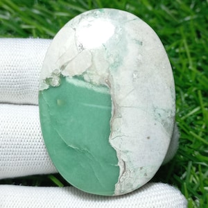 Puede incluir: Gema ovalada con un diseño dividido. Una mitad es de color verde claro sólido, mientras que la otra mitad es una mezcla de blanco y verde claro con una textura veteada. La piedra está pulida y lisa.