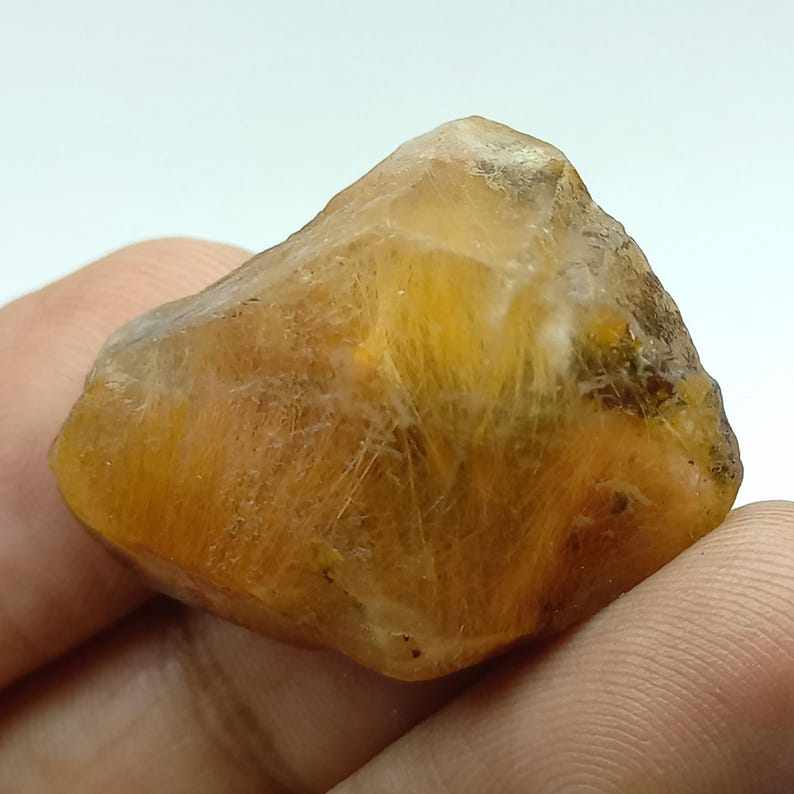 May include: A raw, golden-yellow gemstone with a crystalline structure. The stone has a translucent quality, with visible internal textures and inclusions. The overall shape is irregular, with a slightly rounded top and a textured surface. The colour varies from light to dark yellow.