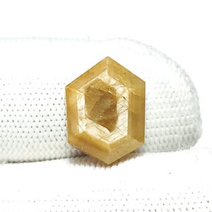 May include: A faceted, hexagonal gemstone with a warm, golden-yellow hue. The gem has a layered appearance with internal inclusions, set against a white, textured background. The cut is geometric and precise.