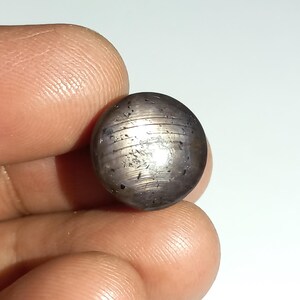 Può includere: Una pietra preziosa rotonda, tagliata a cabochon, di colore grigio con un bagliore argentato. La pietra presenta un motivo striato ed è incastonata in una montatura a castone.