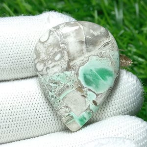 Puede incluir: Una piedra preciosa en forma de lágrima con un patrón de mosaico de tonos blancos, grises y verdes. La piedra presenta una zona central prominente de verde vibrante, rodeada por una matriz de colores más claros. La superficie pulida refleja la luz.