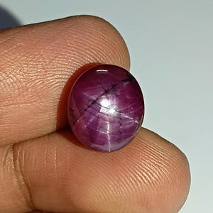 Könnte beinhalten: Ein polierter, ovaler Rubin-Edelstein. Der Edelstein ist ein tiefes Rot mit einem Sechs-Strahl-Stern-Effekt. Die Oberfläche ist glatt und reflektierend, wobei sich das Licht von dem Sternmuster reflektiert.