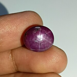 Könnte beinhalten: Ein polierter, runder Rubin-Edelstein im Cabochonschliff. Der Edelstein hat eine tiefe, satte magenta Farbe mit sichtbaren inneren Einschlüssen und einem sternartigen Effekt. Der Stein wird zwischen Fingern gehalten.