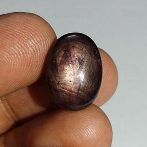 May include: An oval-shaped cabochon gemstone with a deep dark reddish-brown hue. The surface displays a visible star-like optical phenomenon known as asterism, with light reflecting in intersecting lines across the polished, smooth, and rounded dome.