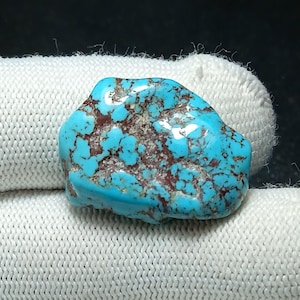 May include: A vibrant turquoise gemstone with a rough, natural texture. The stone features a striking pattern of reddish-brown veins and inclusions throughout its surface. The gemstone is displayed against a white background.