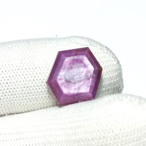 May include: A faceted, hexagonal gemstone with a vibrant pink hue. The gem is held against a white, textured background. The gem's facets catch the light, showcasing its clarity and color. This gemstone is likely used in jewelry.