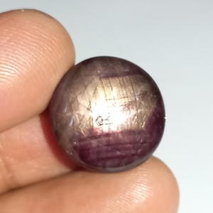 May include: A round, cabochon-cut gemstone with a deep red colour and a silvery sheen. The stone has a star-like pattern on its surface.