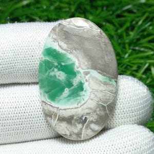 Puede incluir: Una gema ovalada con una mezcla de colores verde, gris y blanco. La piedra tiene una superficie pulida con apariencia veteada. Las áreas verdes tienen una calidad vibrante, casi translúcida, mientras que las secciones grises y blancas son más opacas. Mide unos 4 cm.