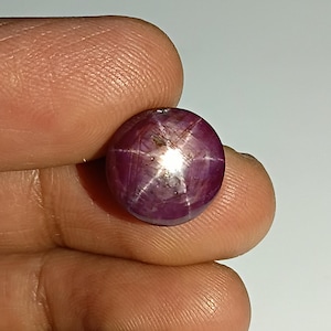 Könnte beinhalten: Ein runder, polierter, tiefroter Edelstein mit einem sechsstrahligen Sternmuster. Der Sterneneffekt entsteht durch Lichtreflexionen von Einschlüssen. Der Edelstein wird zwischen Fingern gehalten, was seine Farbe und seinen Glanz hervorhebt.