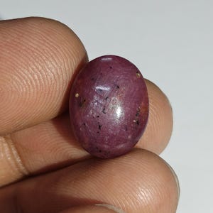 Puede incluir: Gema de rubí pulida, de forma ovalada. La gema es de un color rojo púrpura intenso con pequeñas inclusiones negras y algunas manchas más claras. La piedra se sostiene entre los dedos, mostrando su superficie lisa y reflectante.
