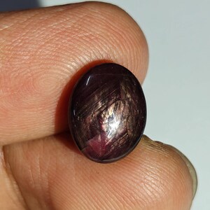 May include: An oval-shaped cabochon gemstone with a deep, dark reddish-purple hue. The surface displays a distinct chatoyant effect, featuring light-reflecting fibrous inclusions that create a shimmering, silky appearance across the polished stone.