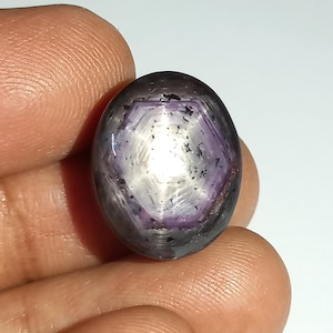 Puede incluir: Una piedra preciosa de zafiro estrella de color púrpura y negro con una estrella de seis puntas en el centro.