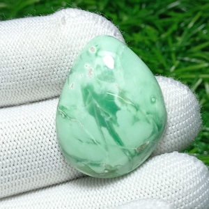 Puede incluir: Una piedra preciosa pulida en forma de lágrima, de color verde claro con patrones blancos arremolinados. La piedra se sostiene en un guante blanco, con un fondo verde borroso.